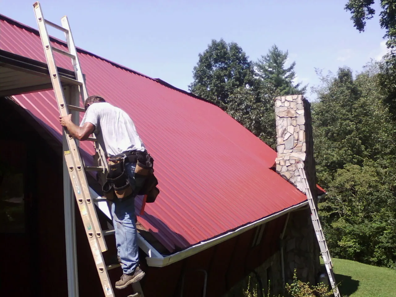 Roofer on a ladder beside a red metal roof and stone chimney — Soffit & Fascia Repair by Roofing Brothers in Waterbury